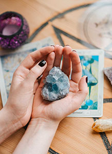 A person holds a blue crystal in their hands, surrounded by tarot cards and a candle on a rustic wooden table.