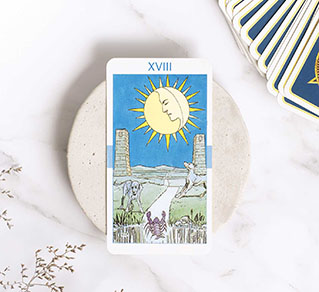 Tarot card XVIII, featuring a sun and moon theme, placed on a stone surface with additional tarot cards in the background.
